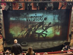The stage with the scrim showing several shadowy figures under the words “The Outsiders”